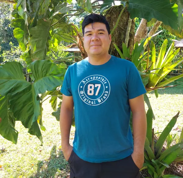 Jim and Gina's son. Smiling man in a teal "Aeropostale 87" t-shirt with hands in pockets, standing in a lush tropical garden.