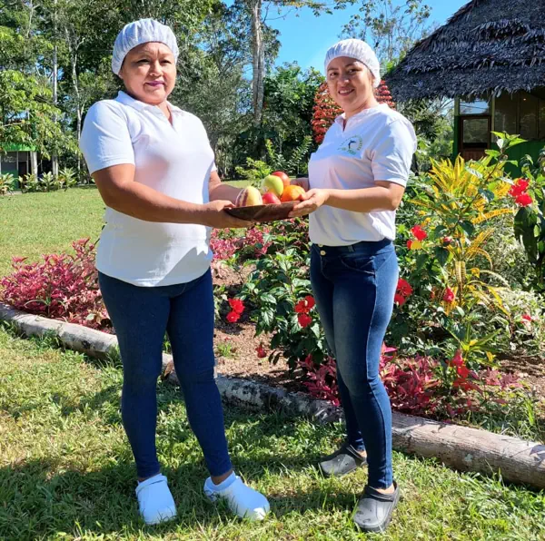 Our cooks. Two women in white shirts and hairnets present a platter of colorful fruit in a sunny tropical garden.