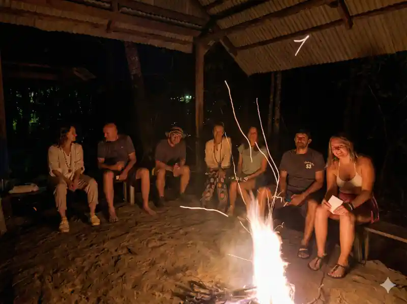 group of men and women sitting around a fire at night, in an outdoor pavillion with simple benches and sandy ground