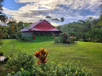 Green building with red roof on a lush lawn, surrounded by vibrant flowers and dense forest under a cloudy sky.