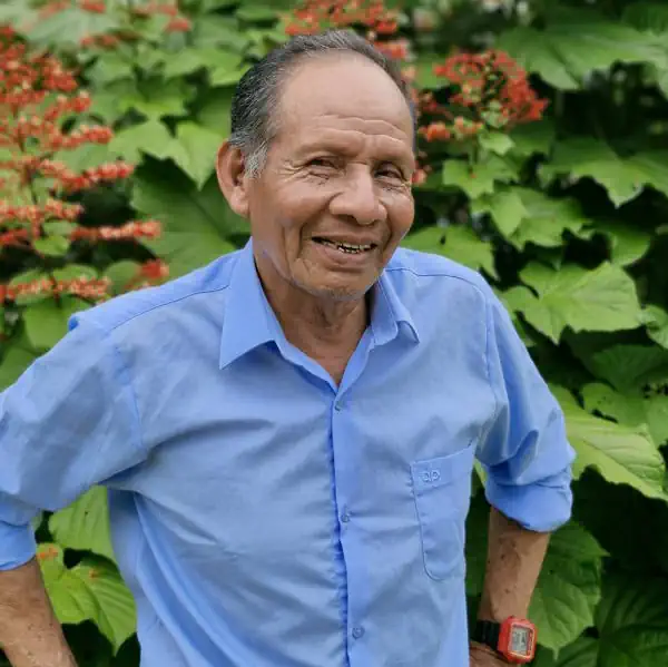 Maestro Don Manain Amacifen. Smiling elderly man in a light blue shirt with a red digital watch, amidst green foliage and red flowers