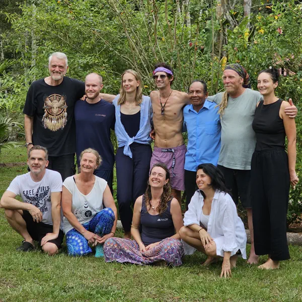 Eleven diverse people, smiling, some standing and some crouching, outdoors on grass with lush green plants.