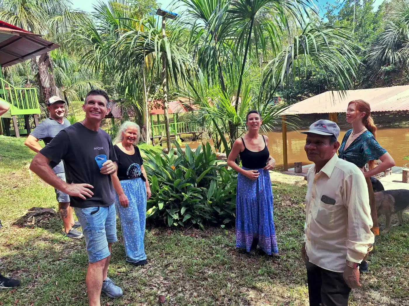 Seven people, including an older man in a cap and a woman in a blue skirt, smiling in a tropical garden with palm trees by a pond.
