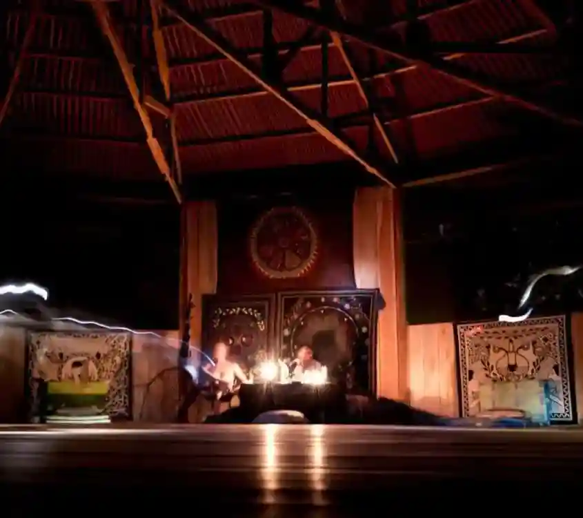 Streaks of light blur movement around two people at a candlelit table, ornate walls, and a thatched roof.