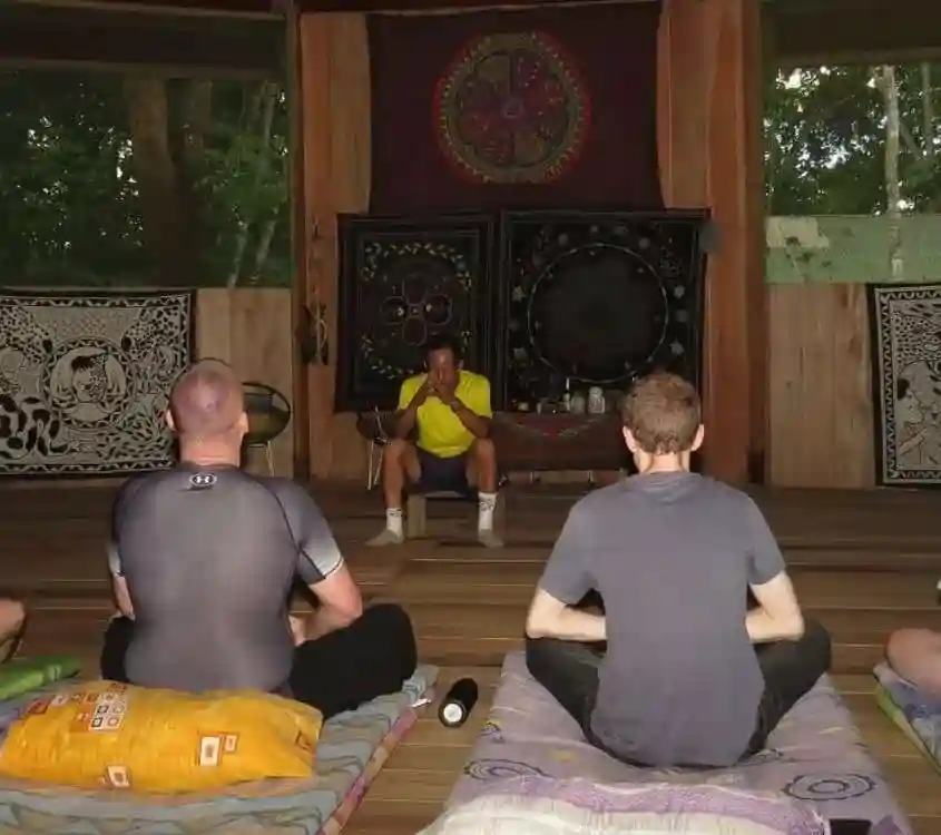 Man in yellow shirt, hands clasped, faces two men meditating with backs to viewer in a room with tapestries.