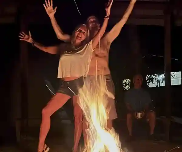 Blurred, joyful figures with raised arms dance around a bonfire at night; a man watches from the dark background.