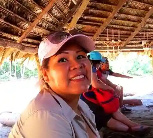 Gina Jim's wife and owner.  Smiling woman in a light cap under a thatched roof, with blurred people sitting behind her.