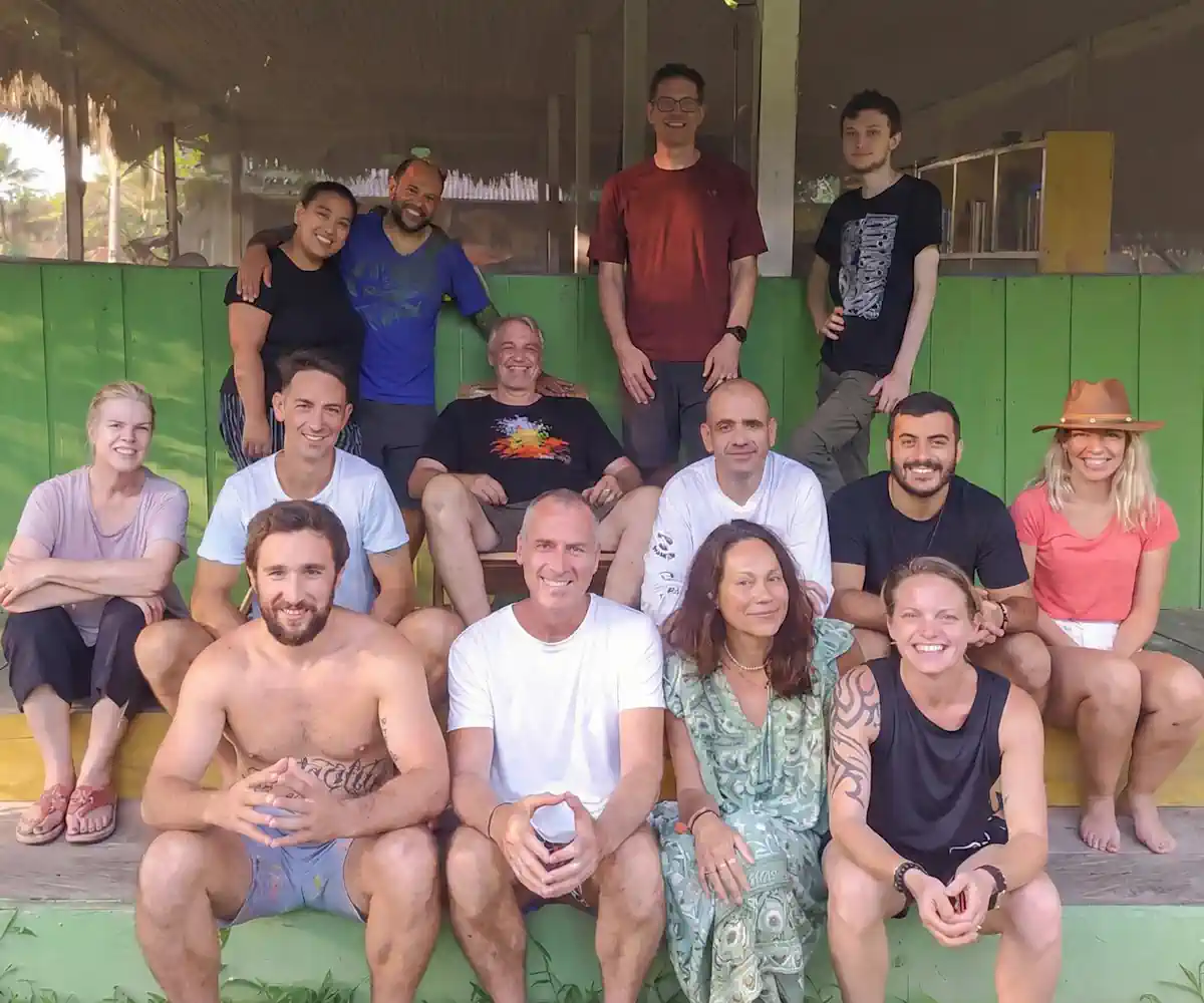 Diverse group of 15 smiling adults, some standing, some sitting on steps outdoors with a green wall.