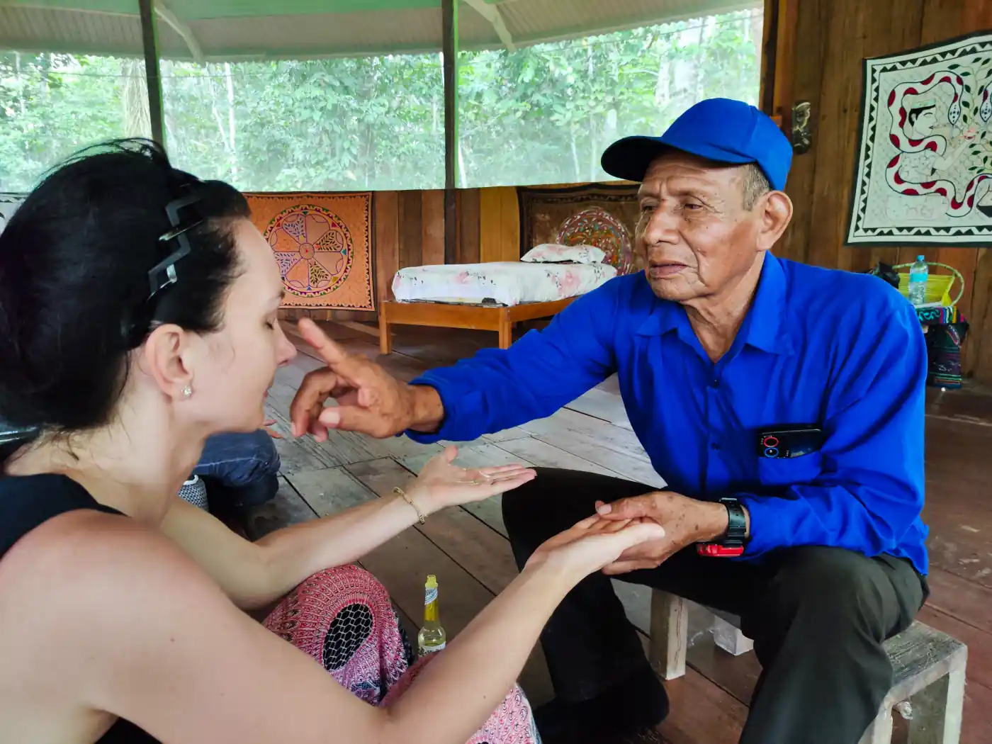 Man in blue cap points at woman's nose, holding her hands in a wooden room with cultural tapestries.