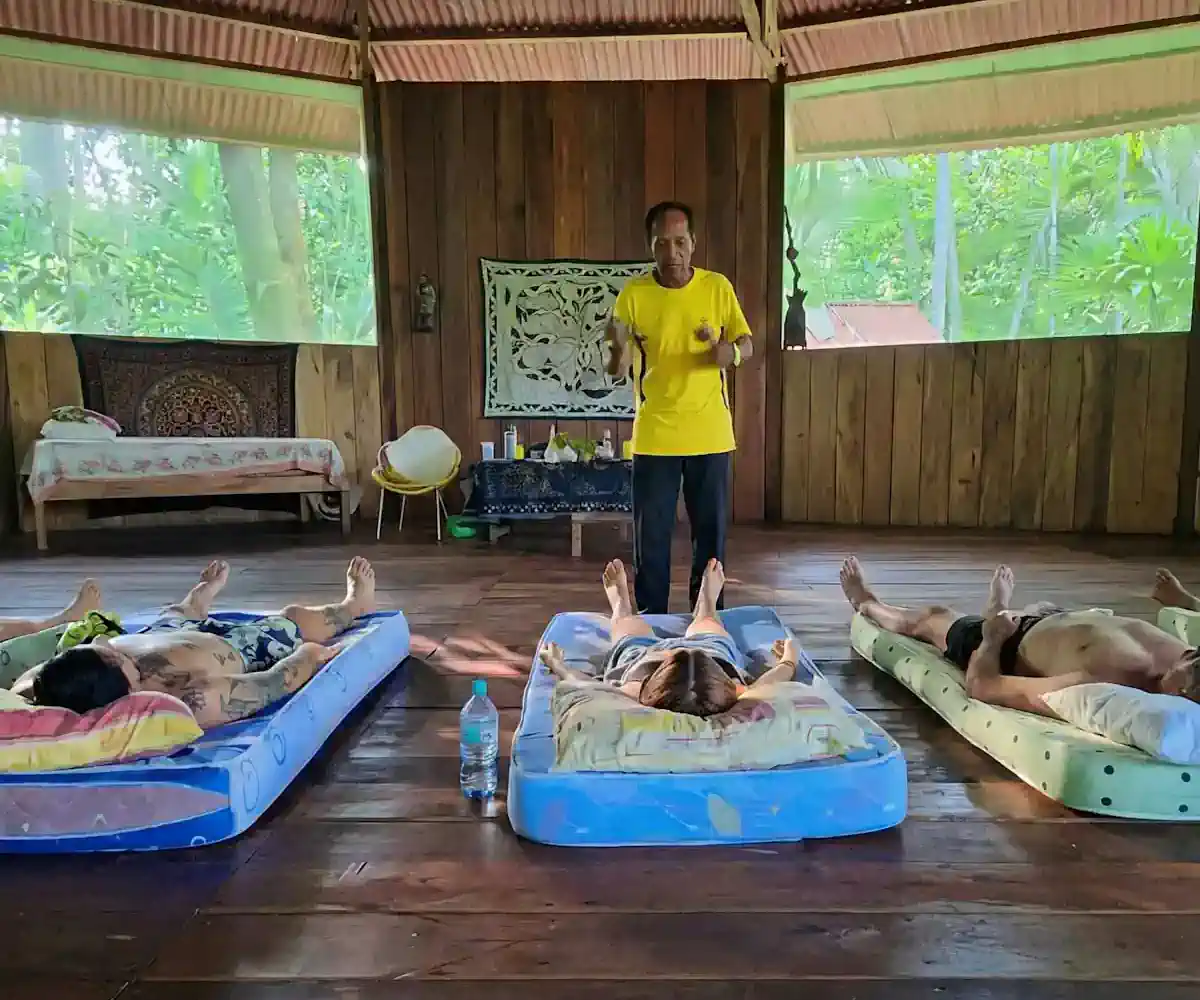 Man in yellow leads a group lying on mats in a rustic, open room with lush green views.