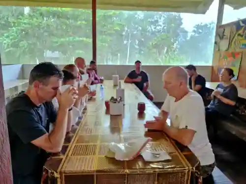 People gathered at a long patterned table, one man sips from a white mug, another gestures, with large windows showing lush greenery