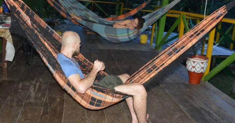 Bald man sits in a plaid hammock on a wooden deck while a woman naps in another behind him