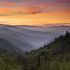 Layers of hazy mountains under a vibrant orange and yellow sunset sky, with dark green forests in the foreground.