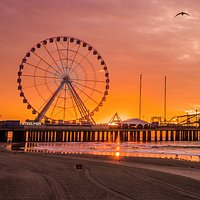 Large Ferris wheel on a pier at sunrise, with an orange sky reflecting on the wet sand and a bird flying.